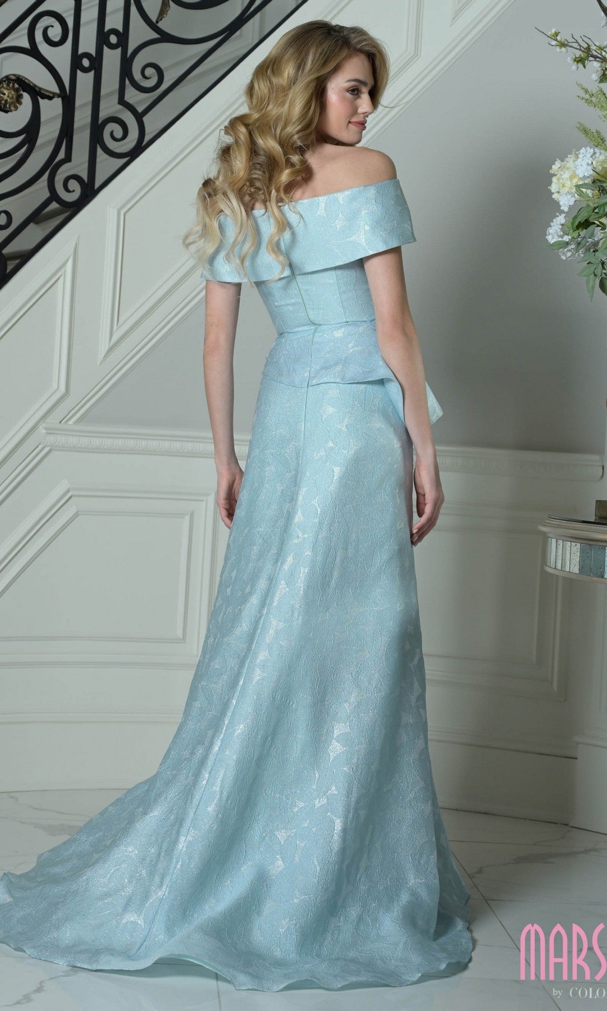 A woman with long wavy blonde hair stands indoors near a staircase, wearing the Marsoni MV1397—a Jacquard long formal dress with an off-the-shoulder neckline and floor-length mermaid silhouette.